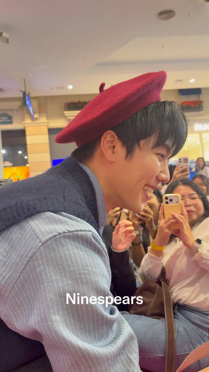 A smiling young man wearing a red beret and a striped shirt sits in front of an audience taking photos; the watermark 'Ninespears' is visible.