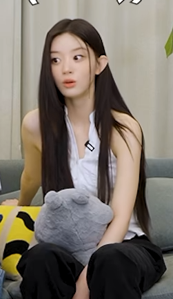 A young woman with long dark hair wearing a white tank top and dark pants, sitting on a gray couch and holding a gray stuffed animal.