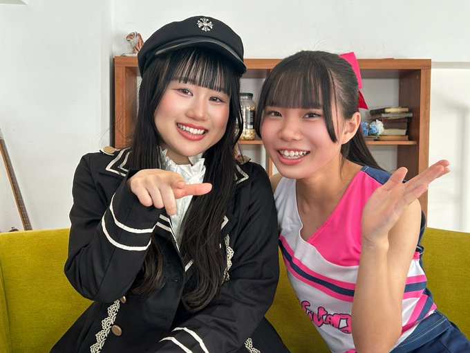 Two smiling young women sitting on a yellow couch; the one on the left wears a black cap and dark jacket, while the one on the right wears a pink bow and a pink/white top.