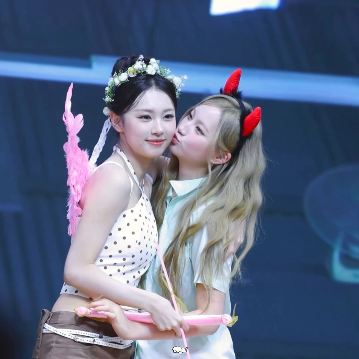 Two performers on stage, one wearing a floral headpiece and pink wings kisses the cheek of the other wearing red devil horns.