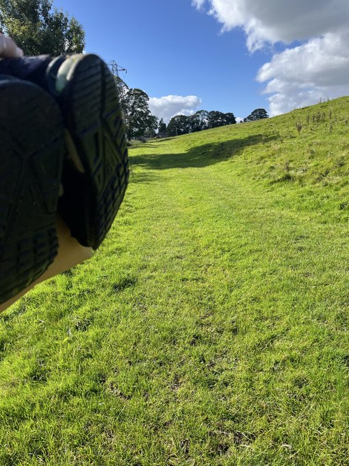 A first-person view of someone relaxing on a grassy hillside under a blue sky with scattered clouds, their sneakers visible in the foreground.