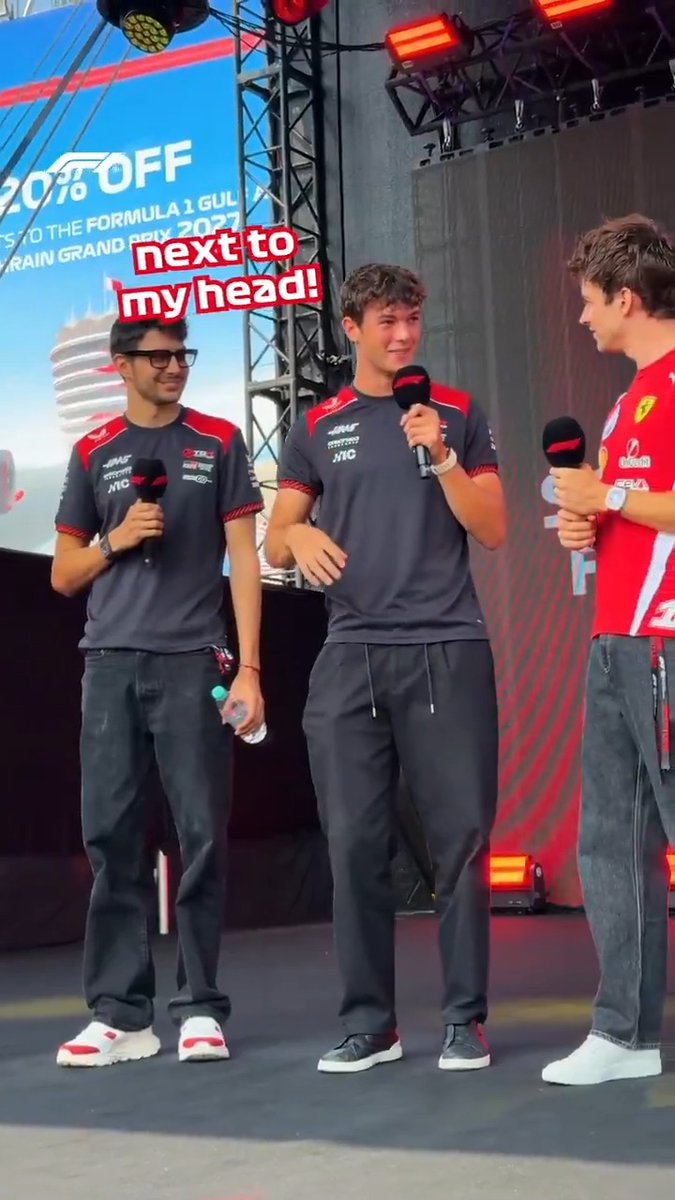 Two F1 drivers in team gear stand on stage at a Bahrain Grand Prix fan event, with one laughing mid-interview as he jokes about a palm-sized spider nearly landing on his head, matching the post's caption.