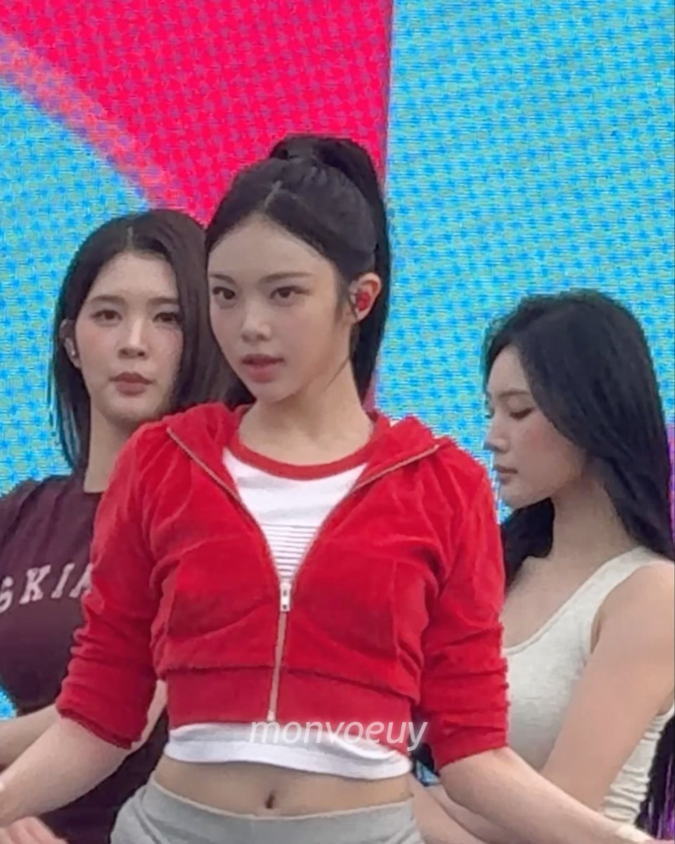 A young woman in a red zip-up jacket over a white top, with her hair in a ponytail, standing with other women against a blue screen background, watermarked "monvoeuy".