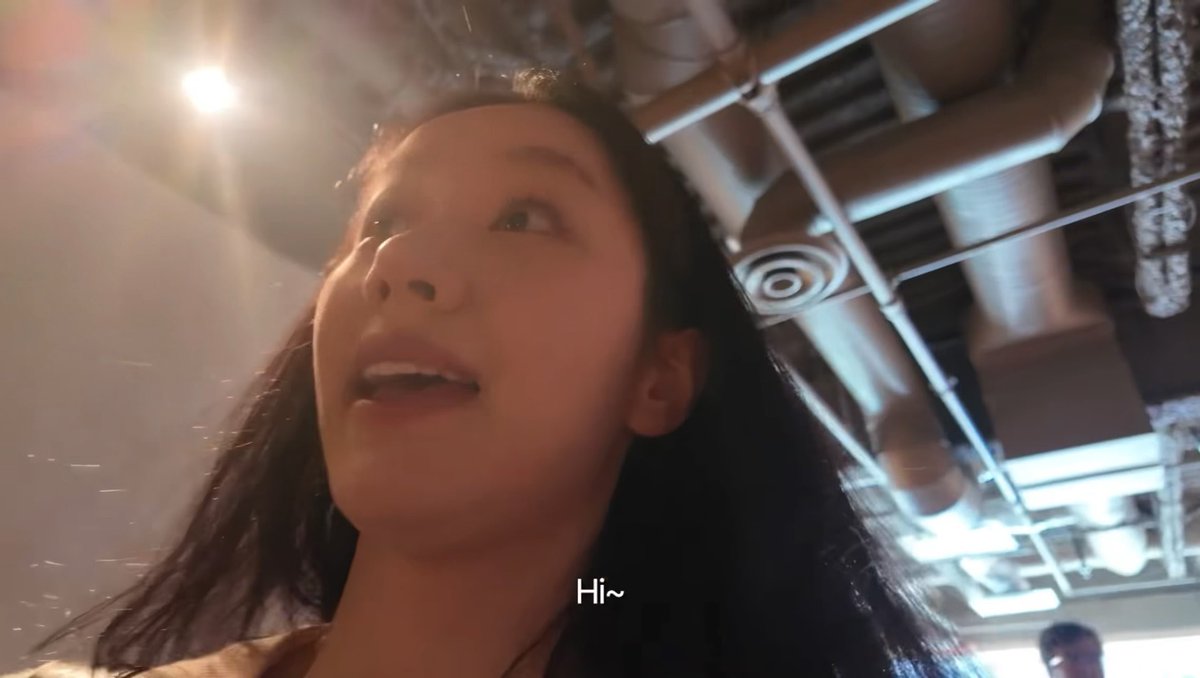 A low-angle close-up of a young woman saying 'Hi~' (subtitled) in what appears to be an indoor venue with exposed ceiling pipes and lighting.