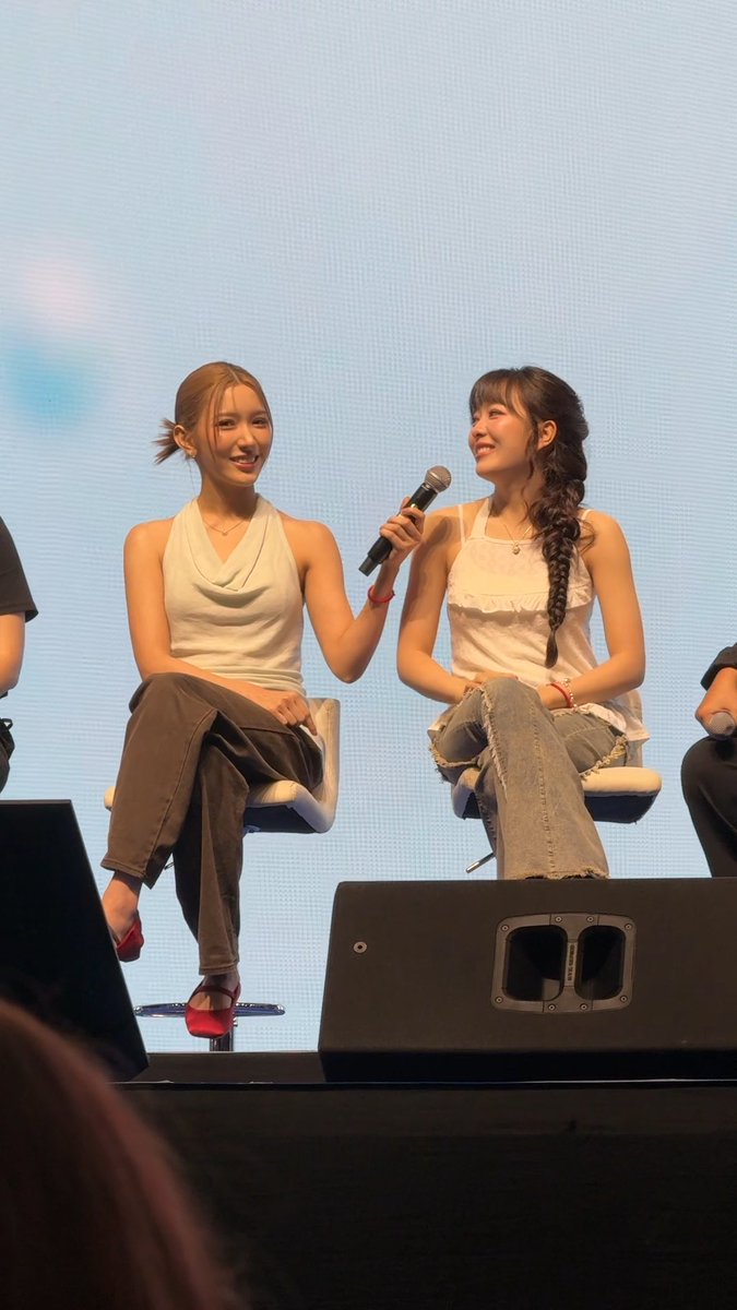 Two young women sit on stage chairs during a panel; one in a white top speaks into a microphone while the other, wearing a white ruffled top and jeans, smiles at her.