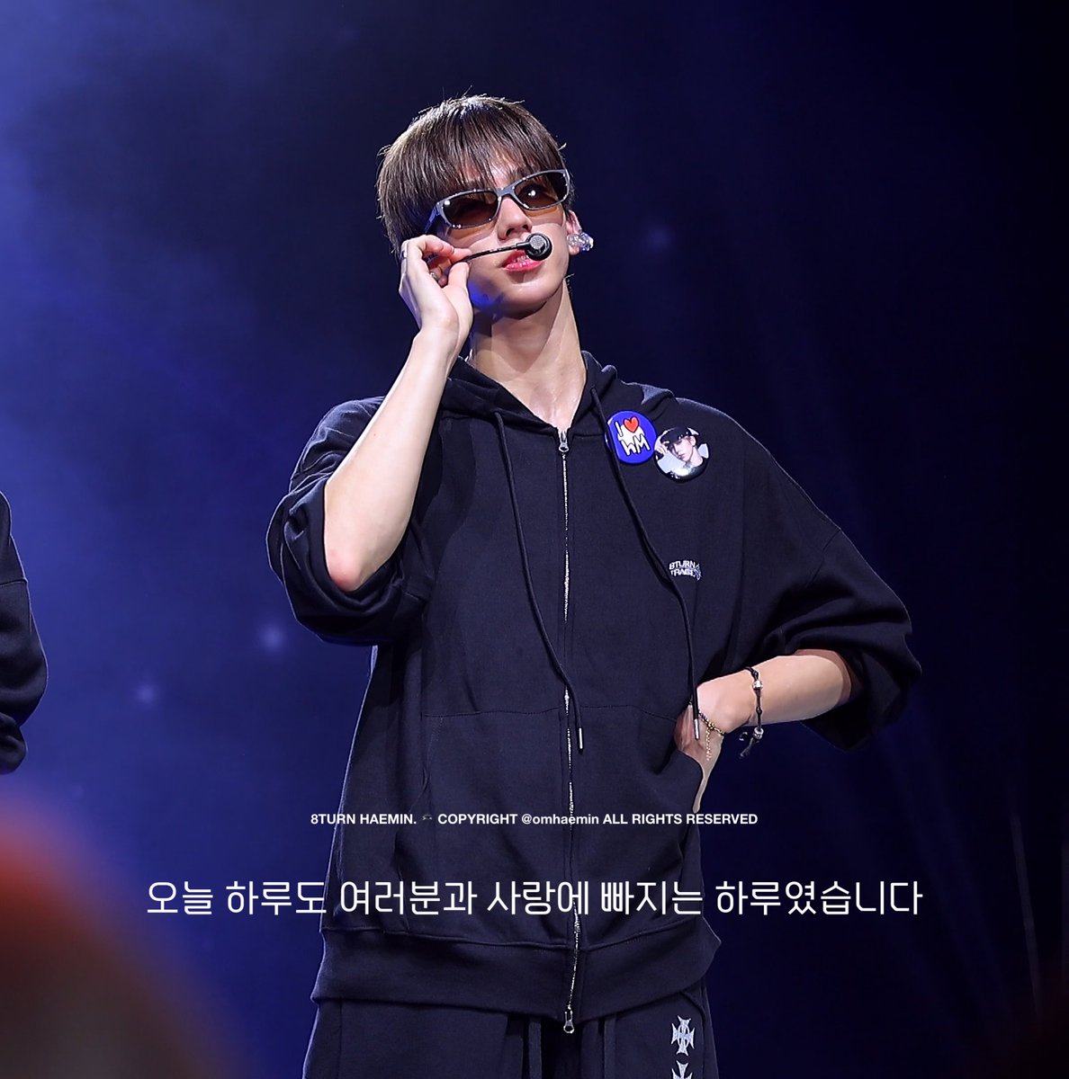 A K-pop performer on stage wears a dark navy zip-up hoodie with 8TURN branded patches, sunglasses, and a headset microphone, with Korean text overlay reading '오늘 하루도 여러분과 사랑에 빠지는 하루였습니다' and watermark '8TURN HAEMIN. COPYRIGHT @onhaemin ALL RIGHTS RESERVED'.
