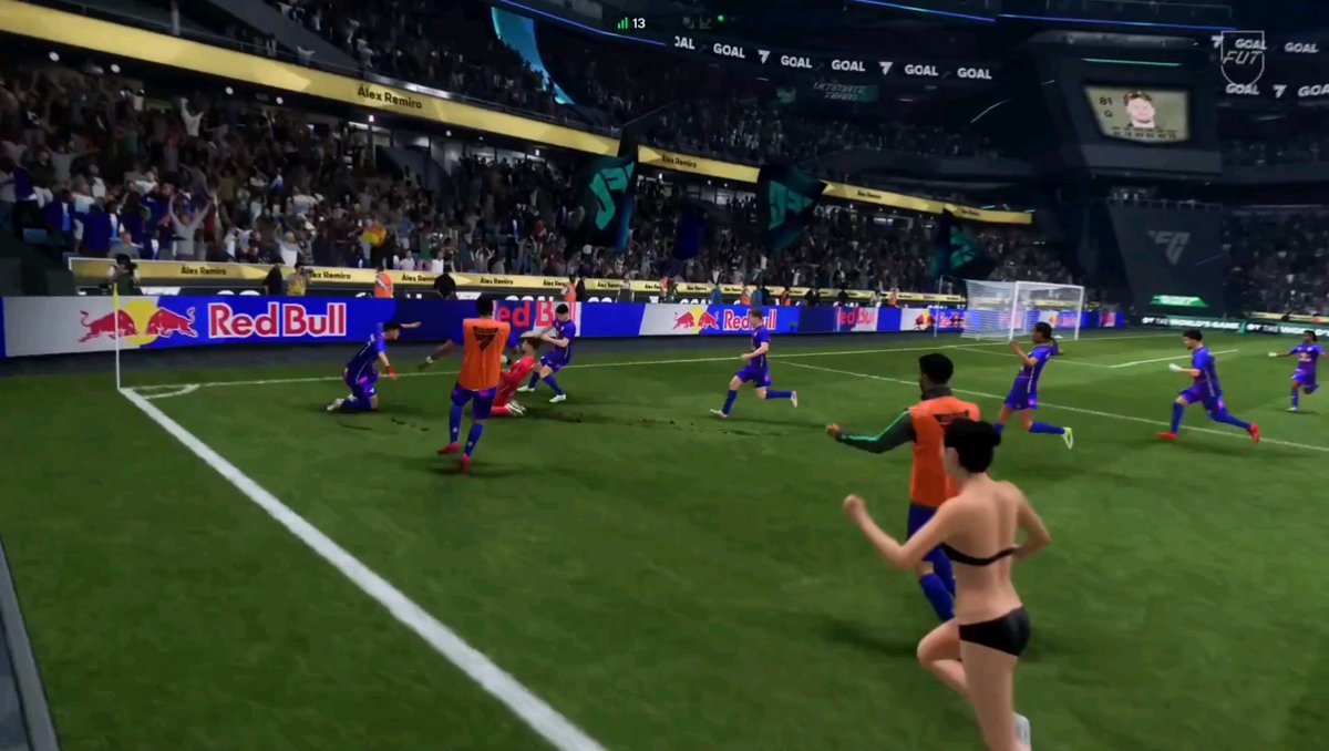 A video game screenshot showing a soccer match in a stadium with Red Bull branding; players in blue and orange uniforms run across the field while a shirtless figure in underwear runs in the foreground.