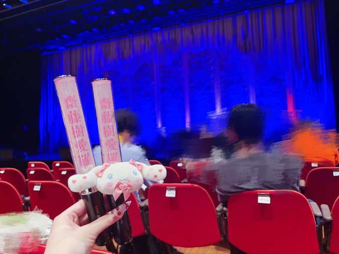 A fan at a concert venue holding two illuminated penlights with Japanese characters (神様関連) and a Cinnamoroll plushie, with red theater seats and a blue-lit stage visible in the background.