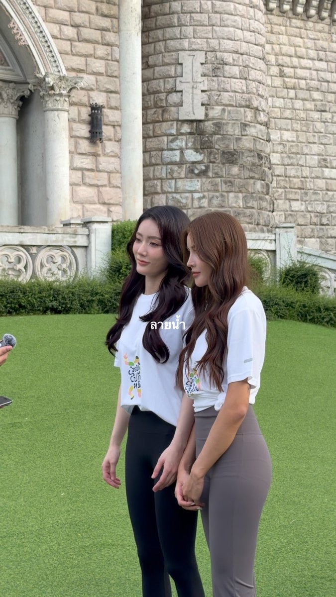 Two young women stand together on a green lawn in front of an ornate stone castle-like building; both wear matching white t-shirts with colorful graphics, and Thai text overlay reads 'ลายน้ำ'.