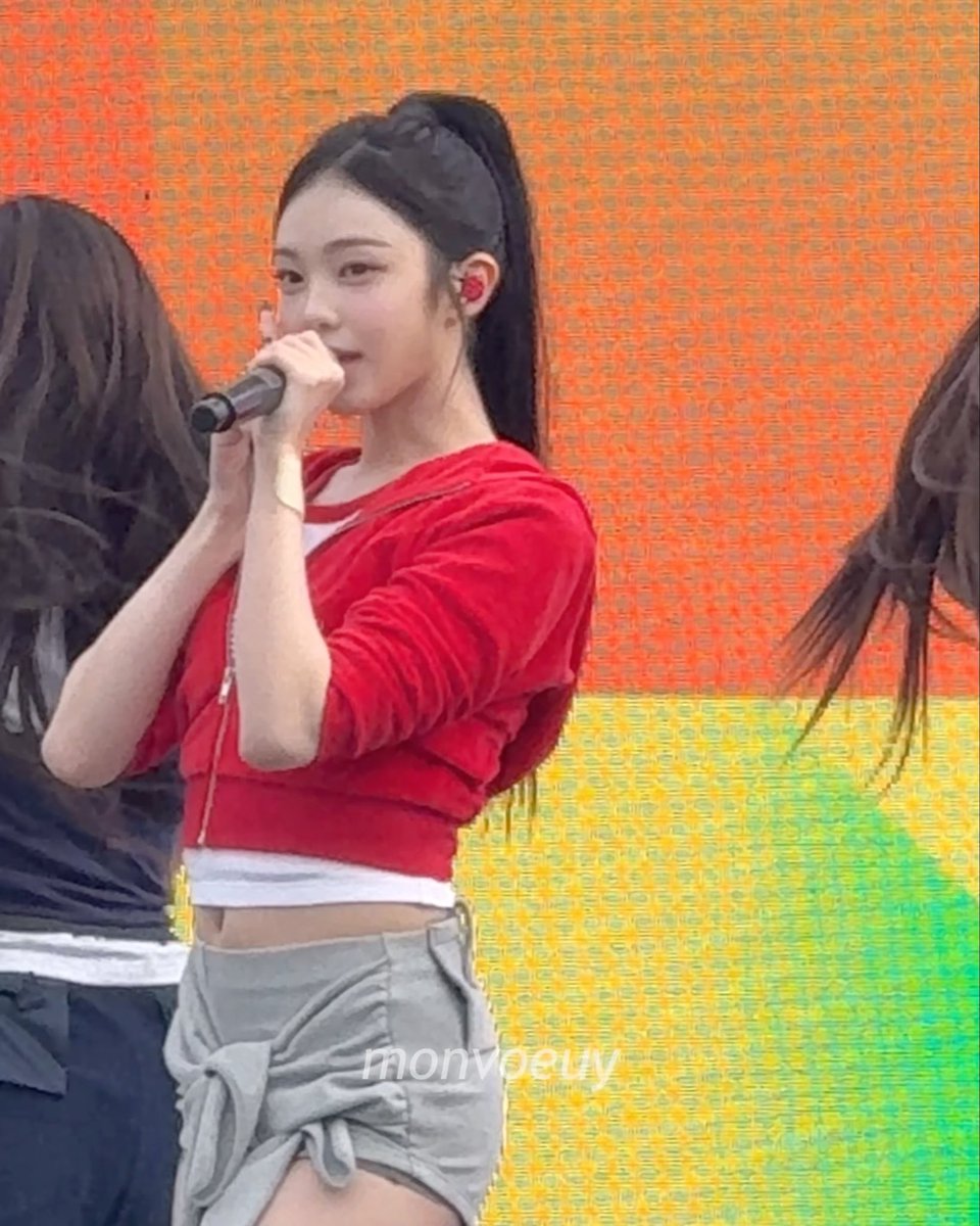 A young woman with long dark hair in a high ponytail sings into a microphone on stage, wearing a red cropped zip-up jacket over a white top, paired with a gray tied shirt around her waist, against a colorful orange and yellow gradient background. A watermark reading 'nonjoeuy' is visible on the image.