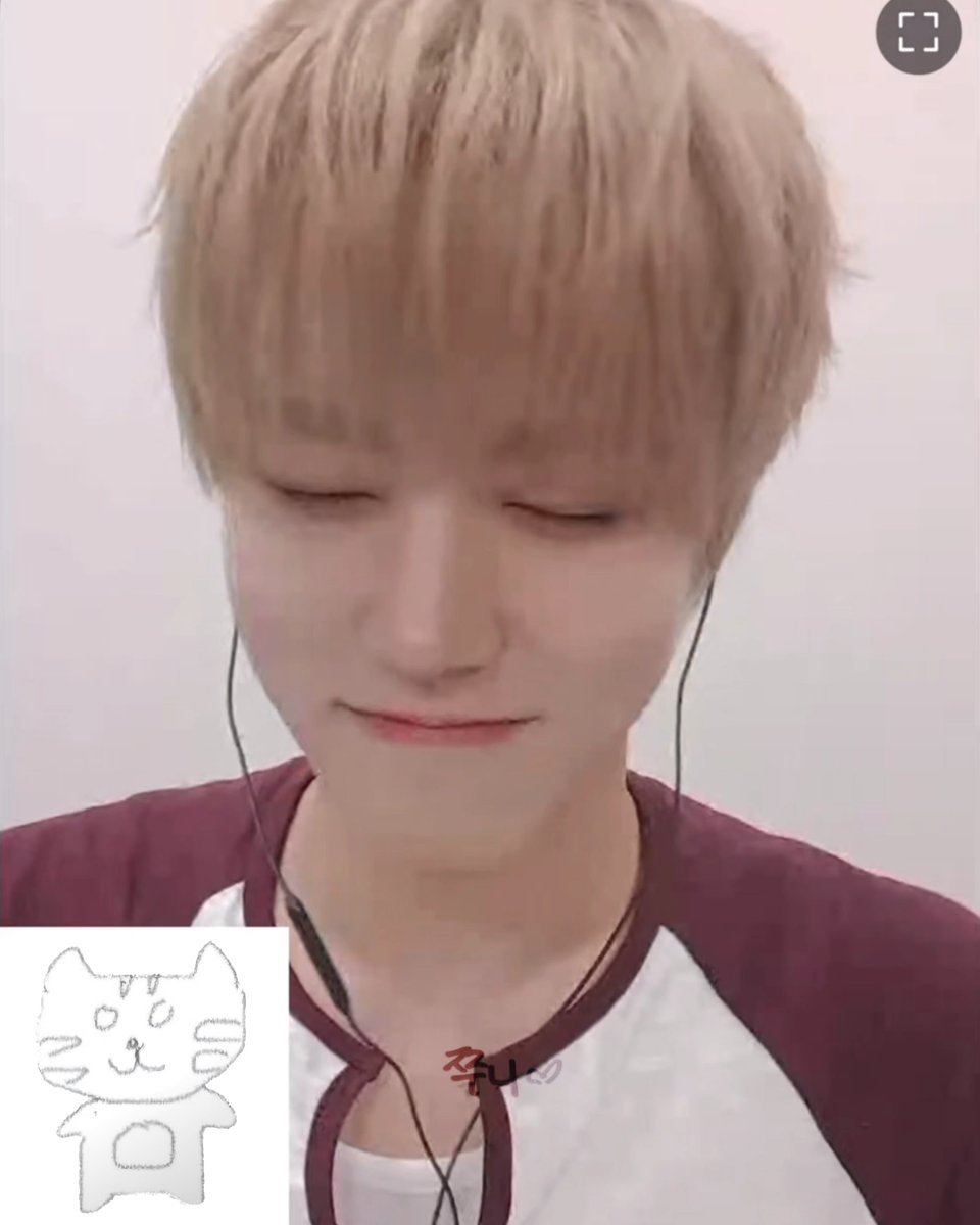 A young man with blonde hair wearing earphones and a maroon-and-white raglan shirt looks down during what appears to be a livestream, with a small cat doodle in the corner and Korean text '주니' visible on screen.
