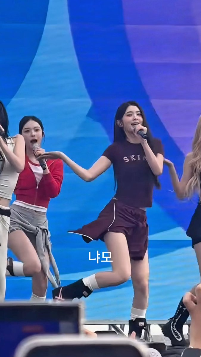 The performer in the center wears a maroon SKIN t-shirt and matching shorts while singing into a microphone on stage, with Korean text '나또' overlaid on the image.