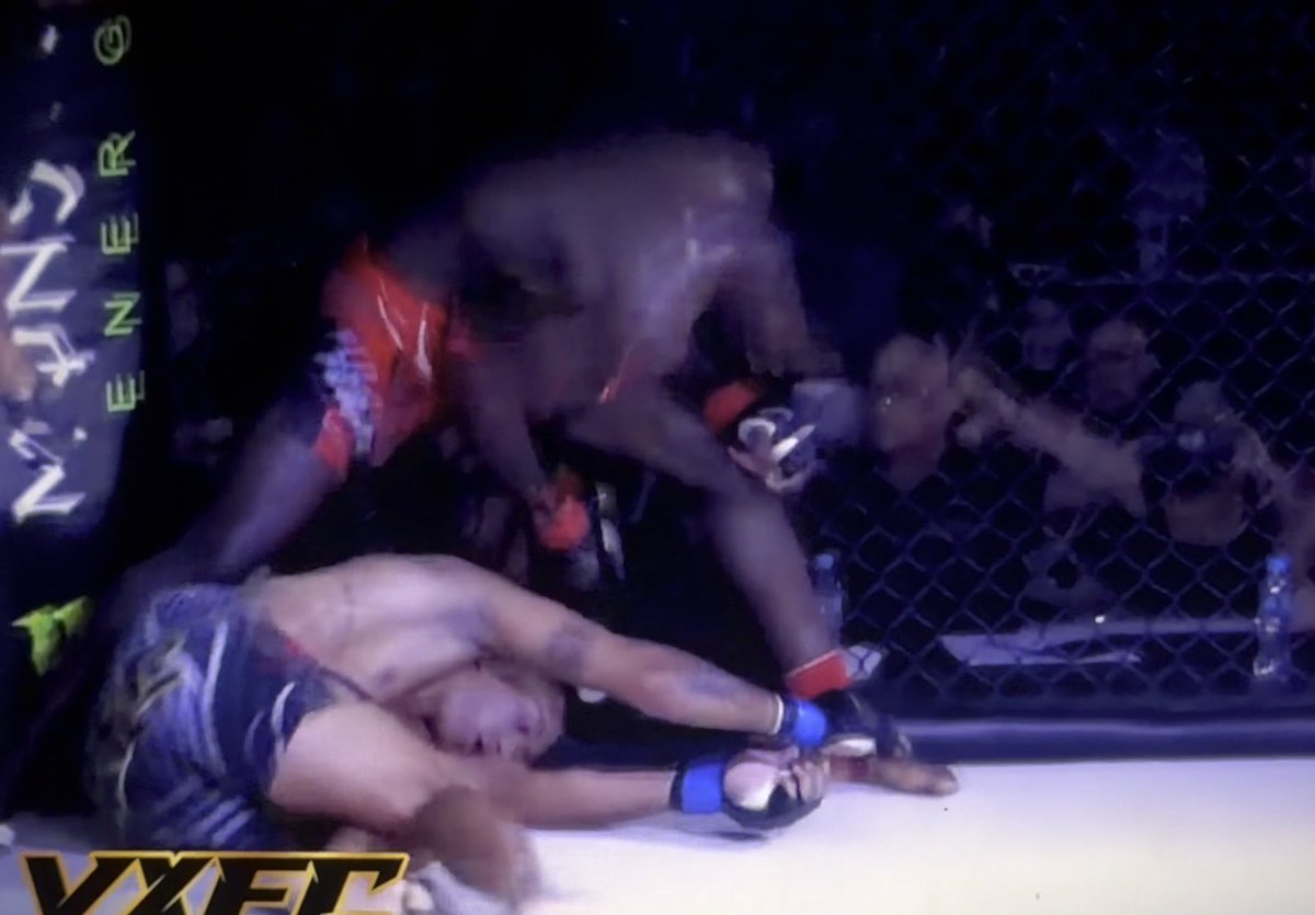 An action shot from a mixed martial arts fight inside a cage, showing a fighter in red shorts delivering a strike to an opponent in dark shorts on the ground.