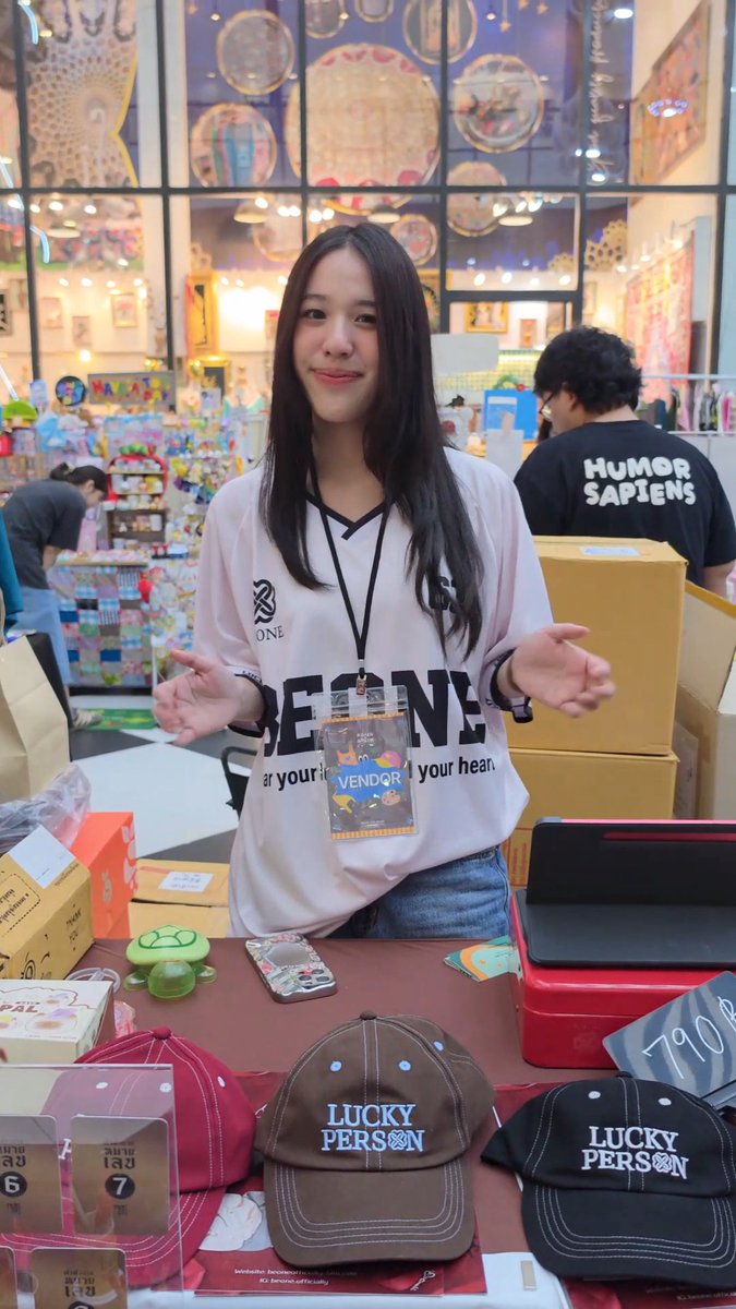 A young woman with long dark hair stands behind a merchandise table at an indoor market, wearing a white jersey with 'BEONE' text and a 'VENDOR' badge, with 'LUCKY PERSON' caps and small turtle figurines displayed in front of her.