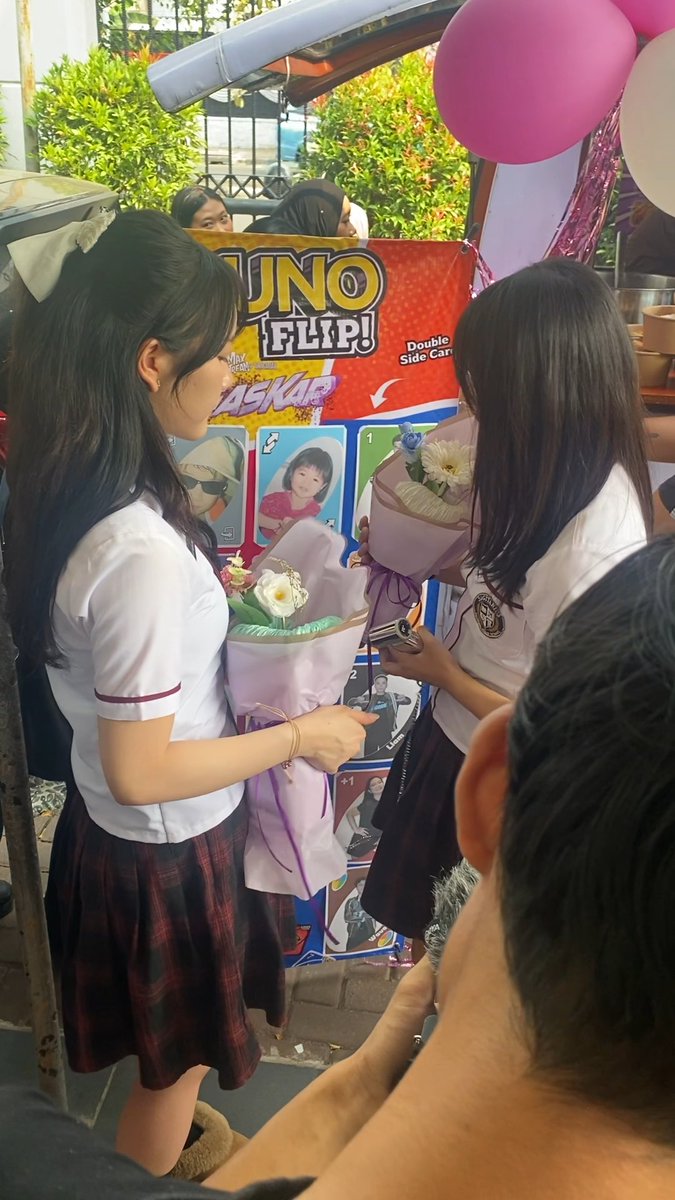 Young women in school uniforms holding bouquets of flowers stand near a carnival-style 'JUNO FLIP!' photo booth decorated with pink and white balloons.