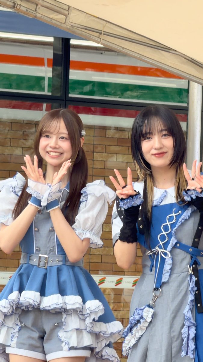 Two smiling young women in elaborate blue and white ruffled idol outfits pose together under a white canopy, both making peace signs with their hands.