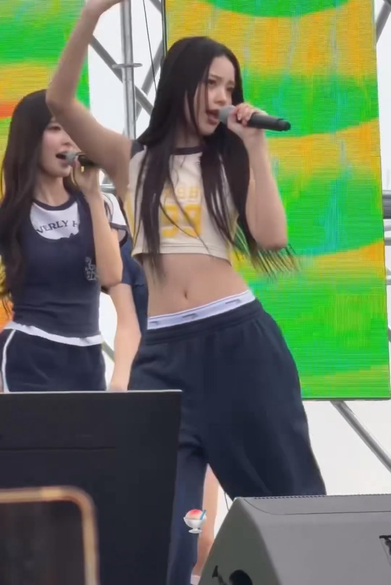 A performer in a cropped yellow and white jersey sings into a microphone on stage, with another singer visible in the background wearing a "BEVERLY H" shirt.