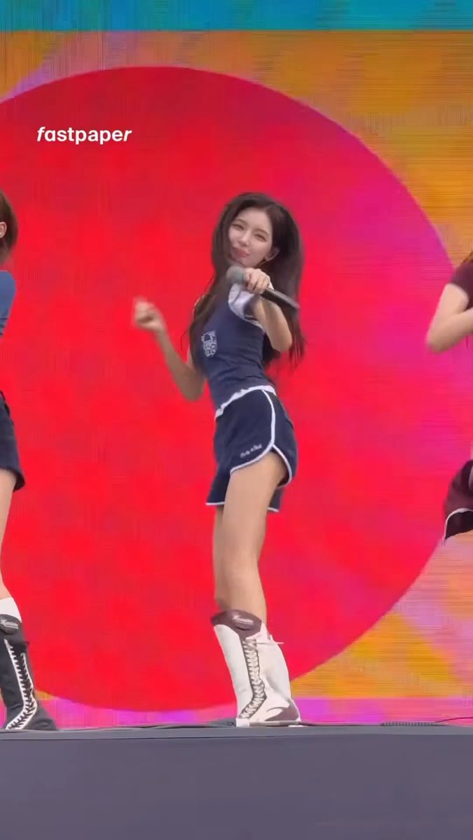 A performer in a navy and white cheerleader-style outfit and tall boots sings on stage with a microphone against a vibrant red and orange background, with the text "fastpaper" visible.