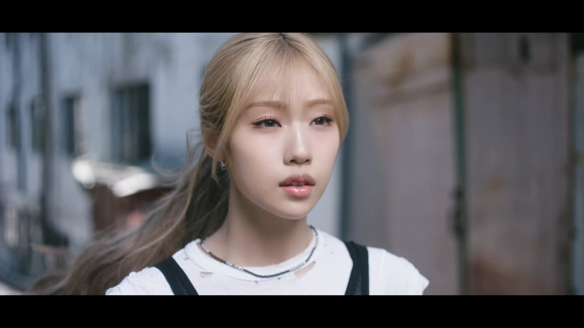 A close-up portrait of a young woman with blonde bangs and dark hair, wearing a white distressed top with black straps and a necklace, photographed with cinematic black bars.