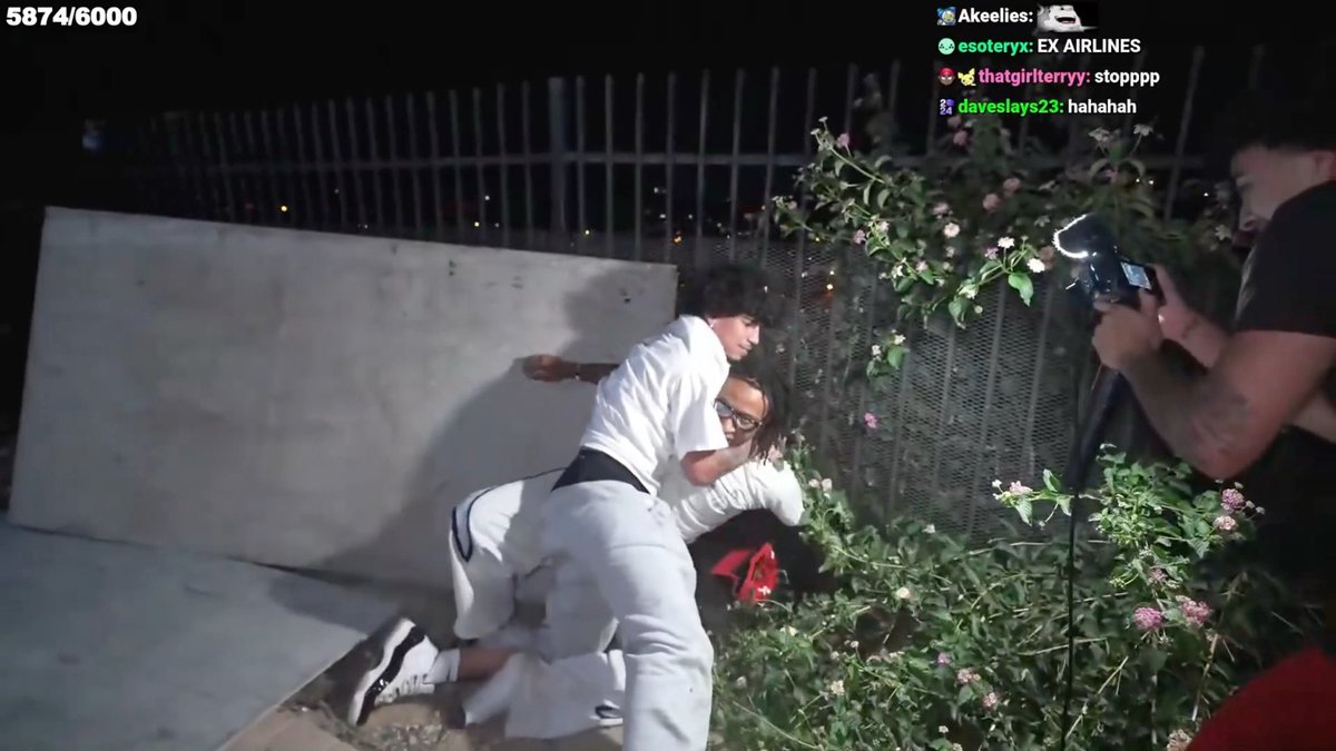 A nighttime livestream screenshot showing two people wrestling on the ground near a fence with flowers, while another person films them with a camera, with chat comments from Akeelies, esoteryx, thatgirlterryy, and daveslays23 visible.