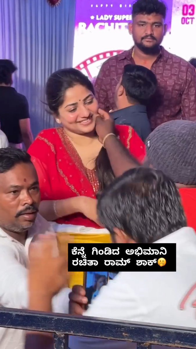 Crowded event scene with a smiling woman in a red traditional outfit holding a child, with a banner reading 'LADY SUPER' visible in background and Kannada text overlay commenting on the situation.