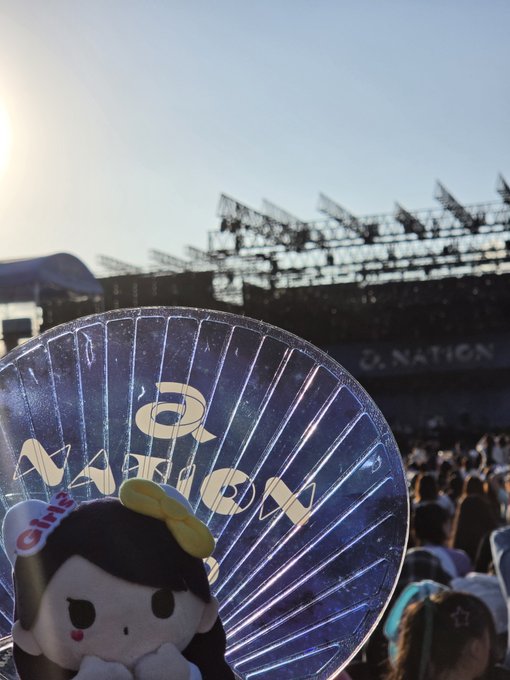 A concert scene with an 'NATION' logo on a handheld fan and stage banner, showing a stuffed toy in the foreground and stadium rigging against a bright sky.