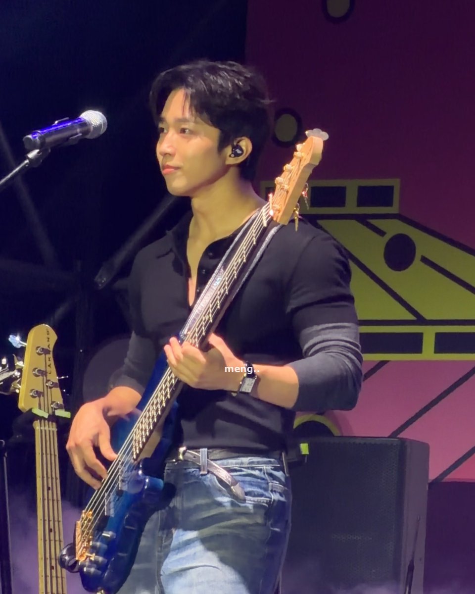 A young male musician performing on stage, wearing a black shirt and jeans while playing a blue bass guitar, with a microphone visible and the watermark 'meng_' on the image.