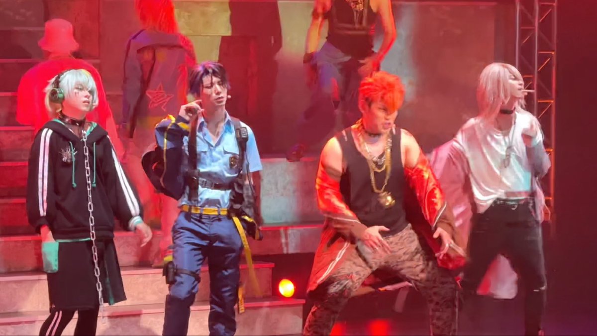 Four male performers on stage under red lights, appearing to be a J-pop or K-pop group performing a choreographed dance routine in colorful, stylized outfits.