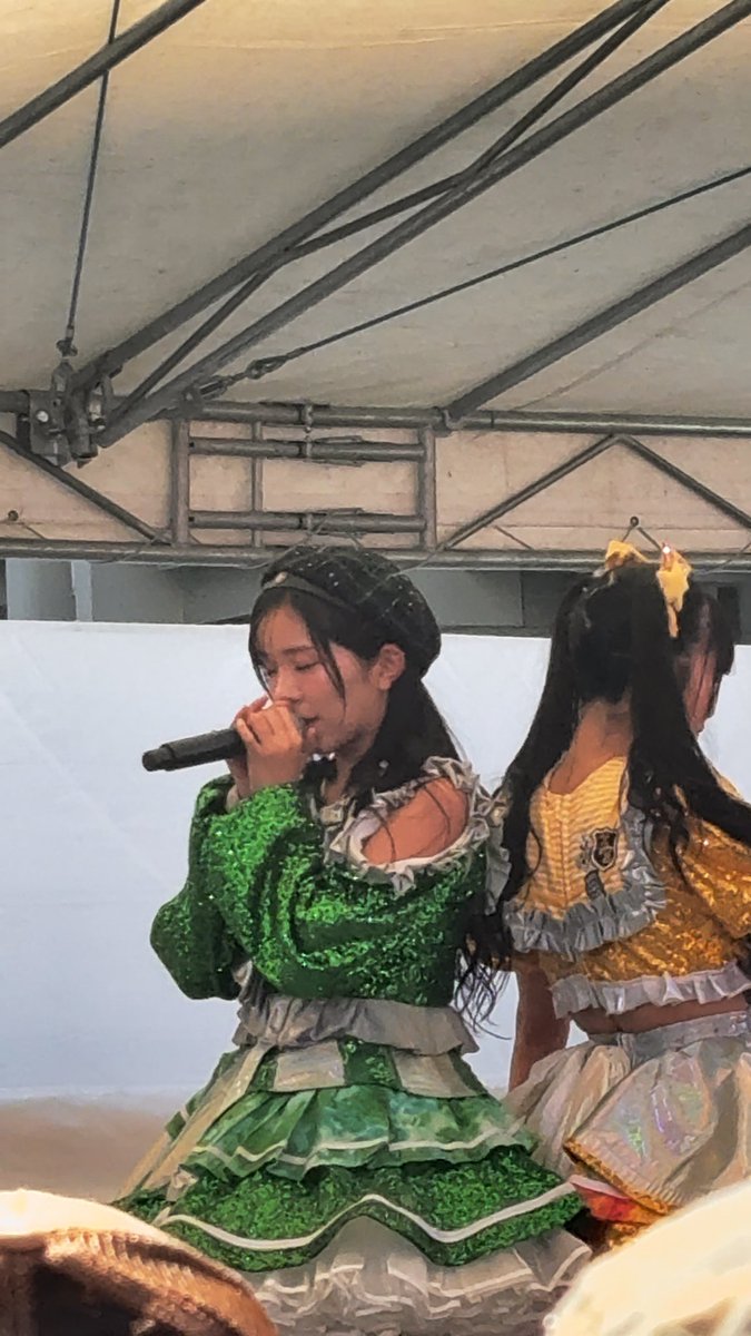Two female performers in ornate green and yellow dresses under a stage canopy, with one singing into a microphone.