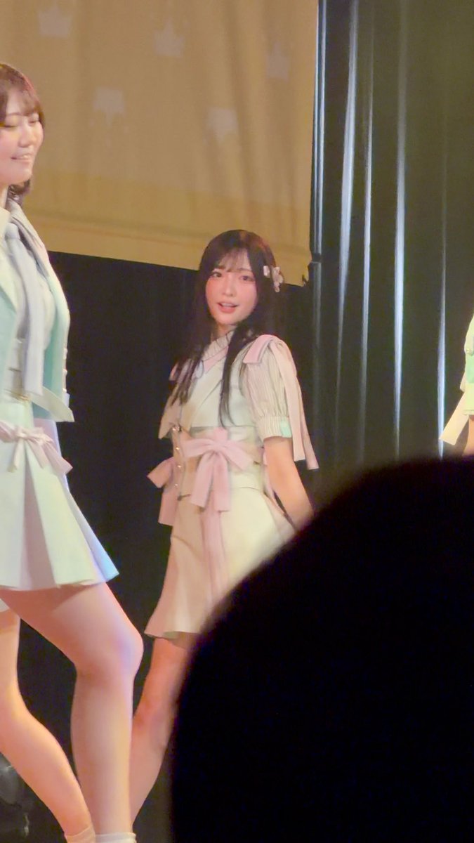 A young Japanese woman in a light green and pink outfit performing on stage, smiling at the camera with a hair accessory.