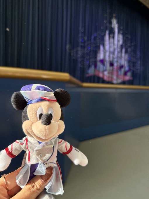 A hand holds a plush Mickey Mouse doll wearing a captain's outfit in front of a Disney-themed backdrop featuring a castle design.