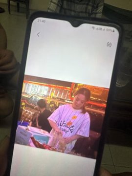 A smartphone displaying a photo of a young woman in a purple 'Vitality' t-shirt sitting at a table.