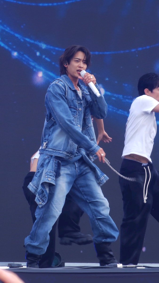 A male performer in a denim jacket and jeans sings into a white microphone on stage, with a large screen displaying blue visuals in the background.