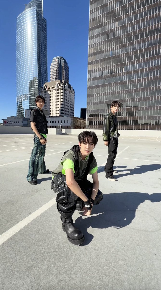 Three young men pose on a rooftop parking lot with modern skyscrapers in the background; one crouches in the foreground wearing black pants and a green-accented top.