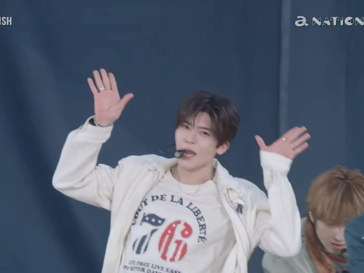 A male performer on tour, identified by the 'NATION' logo, sings passionately while wearing a white jacket and a '76' t-shirt, with both hands raised.