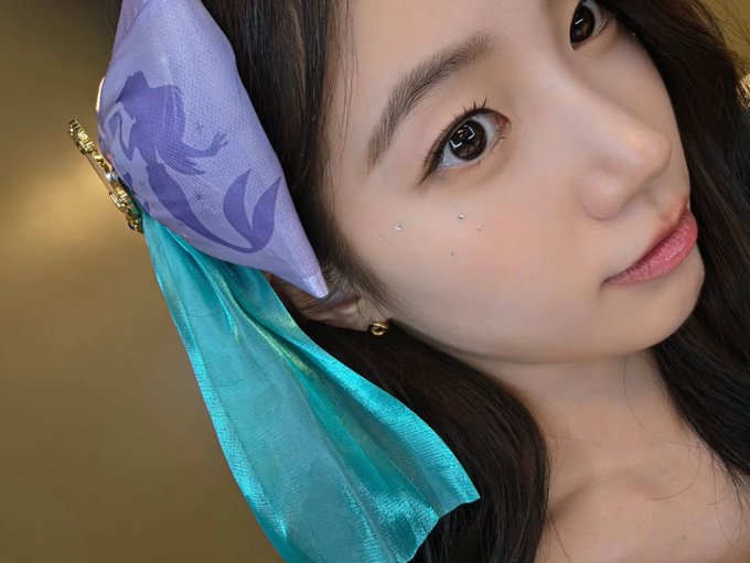 A close-up portrait of a young Asian woman with long dark hair, wearing a purple floral hair accessory and teal scarf, with small rhinestones decorating under her eye.