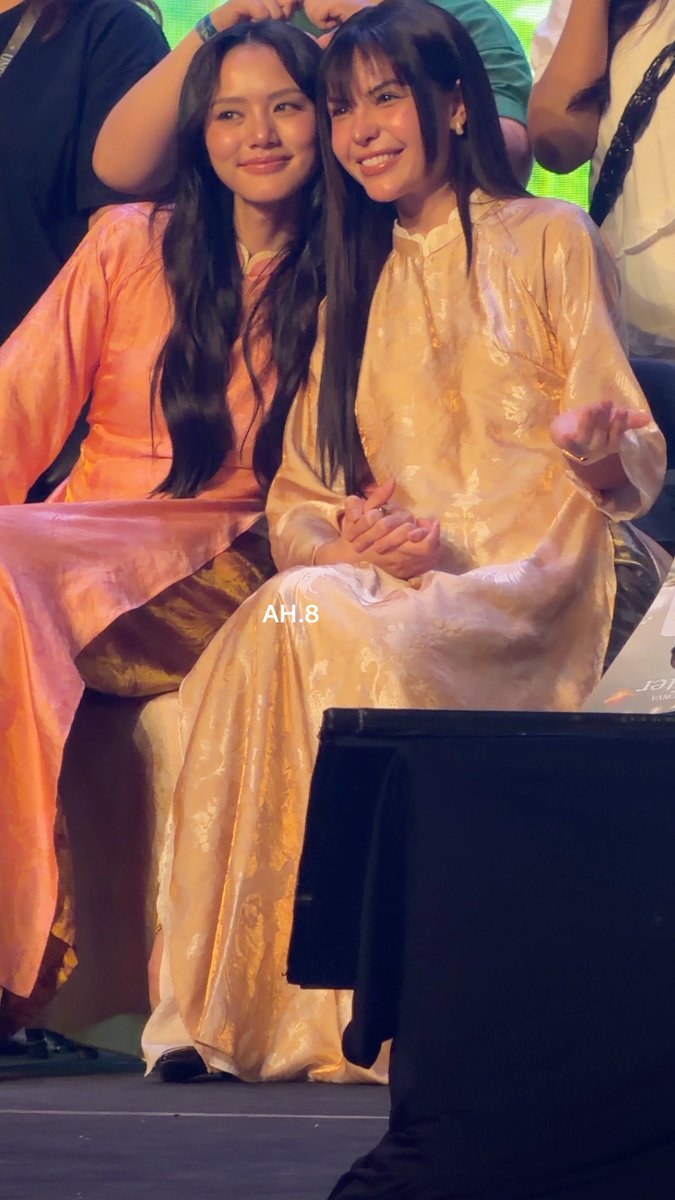 Two women wearing traditional silk áo dài in peach and cream-gold tones sit closely together, smiling warmly as they hold hands, with the watermark "AH.8" visible across the image—capturing an intimate fan moment that complements the affectionate hashtags like #LMSYUnfoldInHCMCity and #LMlookmhee referencing the popular Thai celebrity duo.