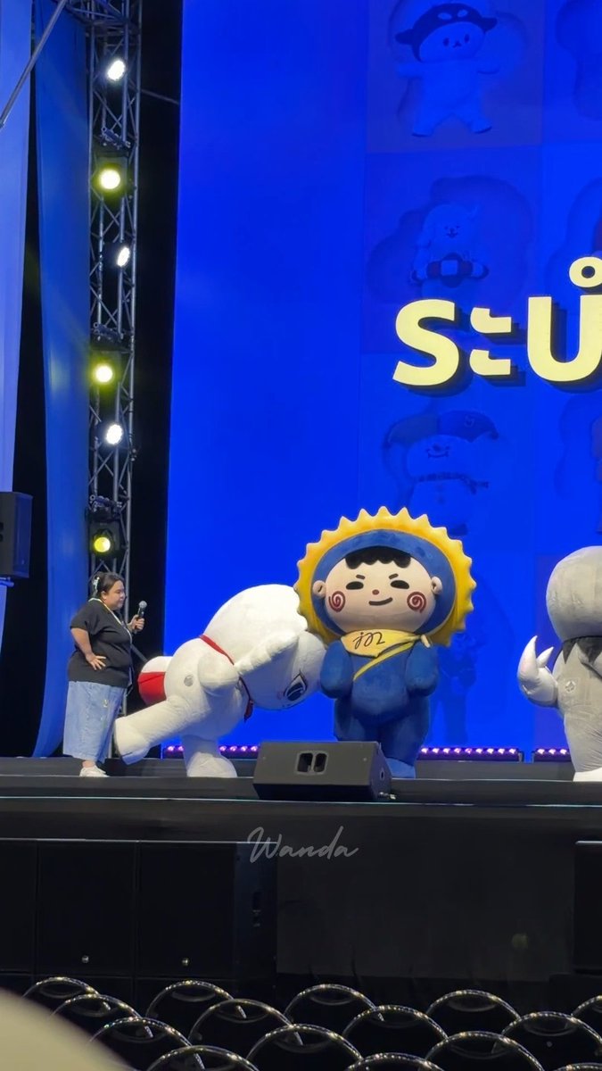 A stage event featuring mascot characters, including one dressed in a blue sunflower costume and another large white character, with a person holding a microphone on the left, against a blue backdrop with mascot illustrations.