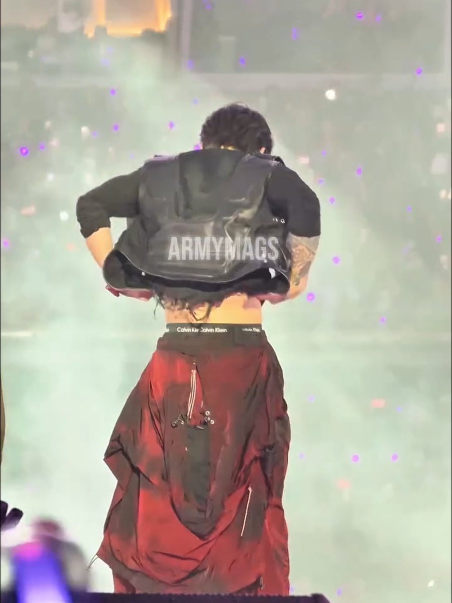 A performer on stage lifts up a black jacket to reveal oversized burgundy Calvin Klein pants, humorously fumbling with the waistband in a moment that perfectly matches the caption "jungkook trying to figure out how his pants work."