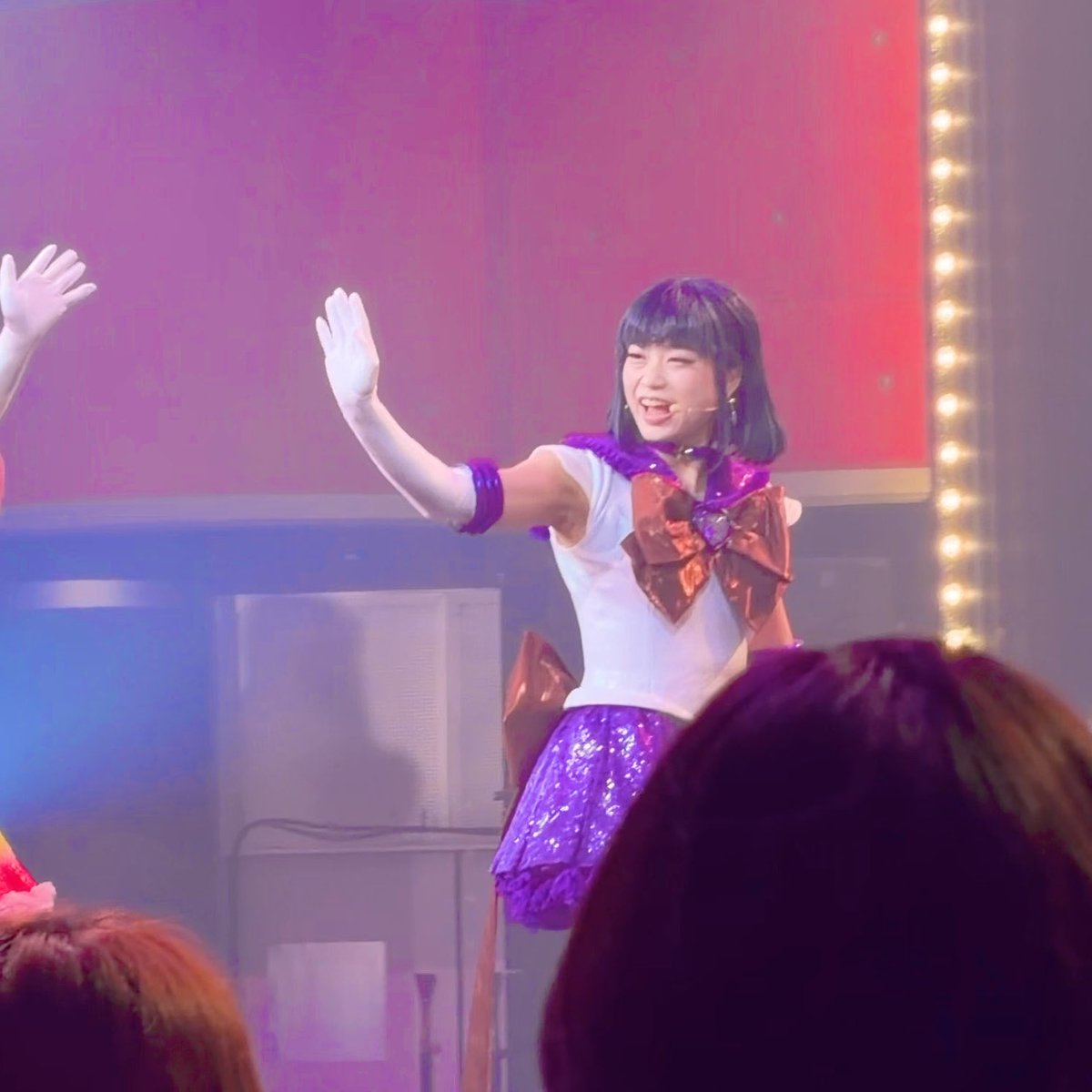 A performer on stage in a white and purple costume with a large orange bow waves to the audience with white gloves, set against a backdrop with stage lights and red/orange lighting.