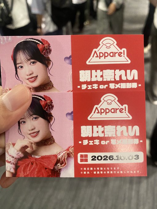 A hand holding two identical promotional cards or tickets with a pink background, featuring a young woman in a red costume, the text 'Appare!', Japanese text '朝比奈れぃ', and a date '2026.10.03'.
