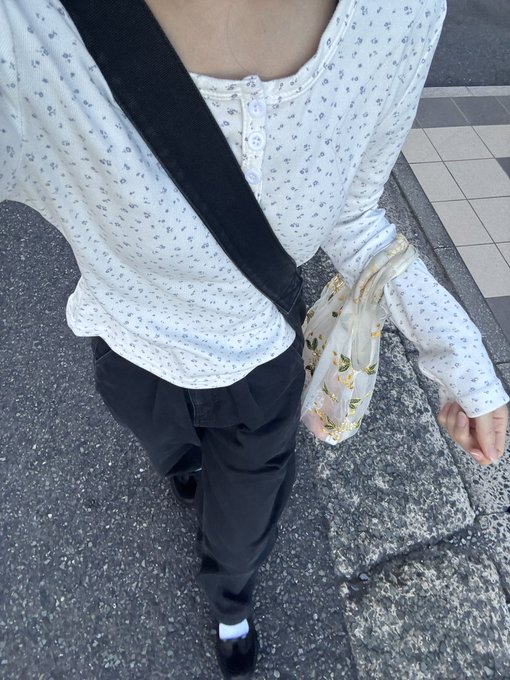 A street style photo showing a person wearing a white floral-print long-sleeve henley top, black pants, and a black crossbody strap, carrying a small cream-colored floral shoulder bag.