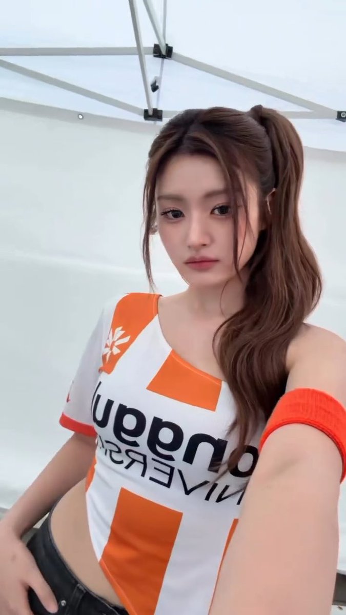 A woman wearing an orange and white 'Uppdom' athletic jersey takes a selfie under a white canopy.