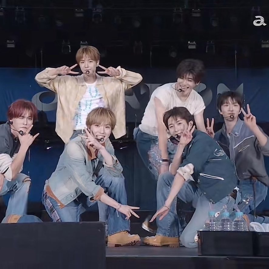 Seven young men pose together on stage, making peace signs and playful gestures while wearing casual denim and layered outfits, with a stylized "a" logo visible in the upper right corner. The image complements the post's caption about Sakuya initiating group photos, showing the members' natural camaraderie and readiness to follow her lead into a coordinated pose.