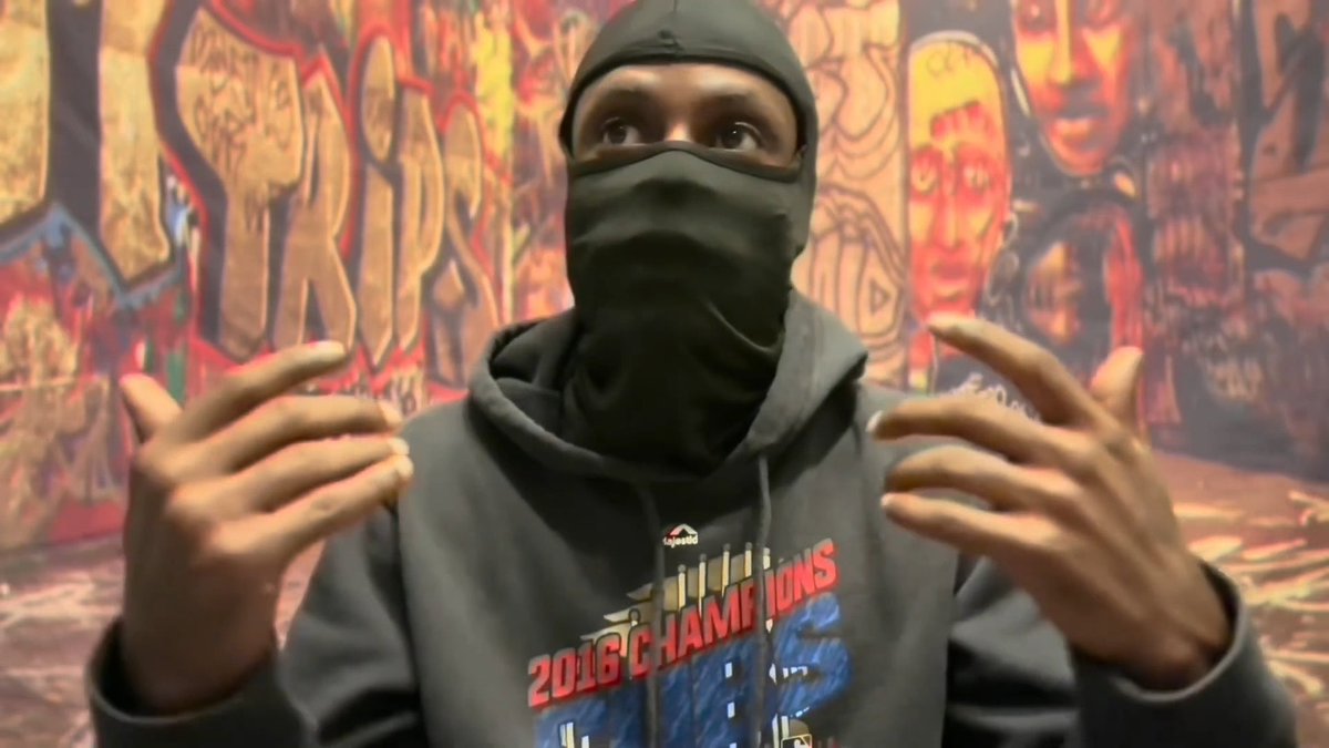 A man wearing a dark hoodie and a face covering (balaclava) standing in front of a graffiti-covered wall, gesturing with his hands. The hoodie reads '2016 CHAMPIONS'.