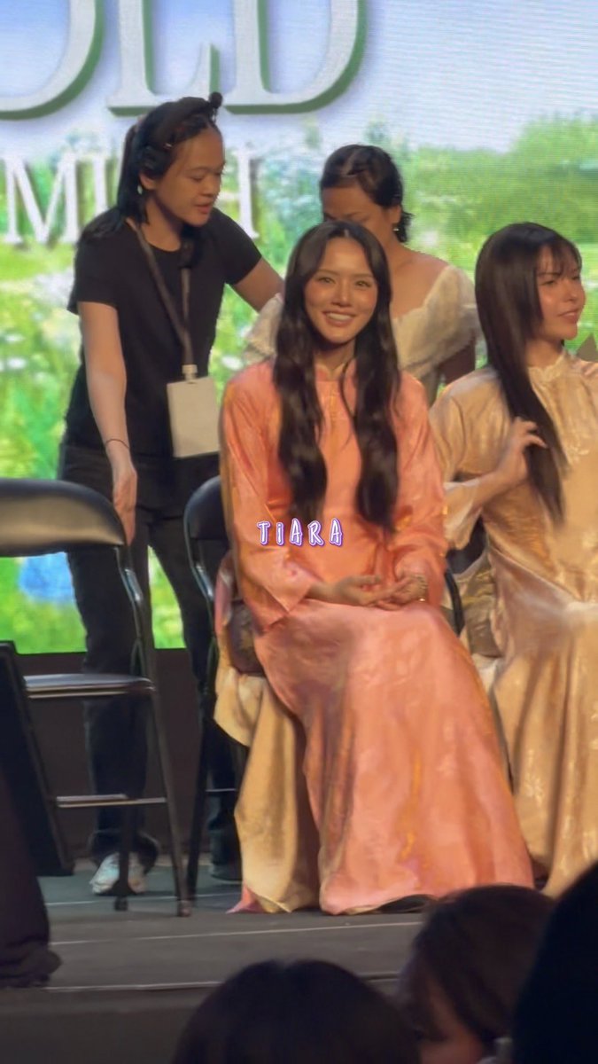 A young woman with long dark hair wearing a peach/orange traditional-style dress sits smiling on stage, with another woman in black behind her; the text 'TIARA' appears in pink/blue letters.