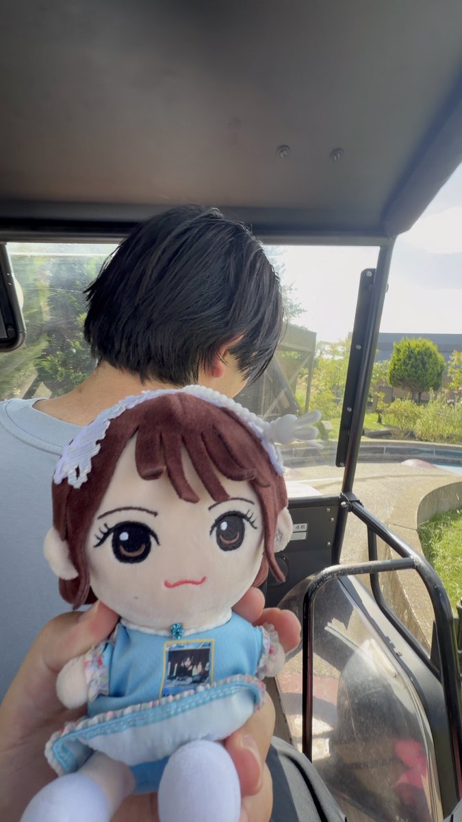 A person with short dark hair wearing a blue shirt sits in an open-air vehicle, while a hand in the foreground holds up a small plush doll of a girl in a blue dress with white lace trim.
