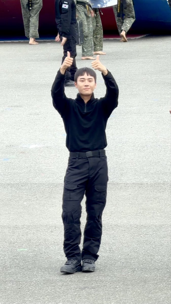 A young man dressed entirely in black, including a long-sleeved shirt and cargo pants, stands outdoors giving a double thumbs-up gesture, with other figures in military-style clothing visible in the background.