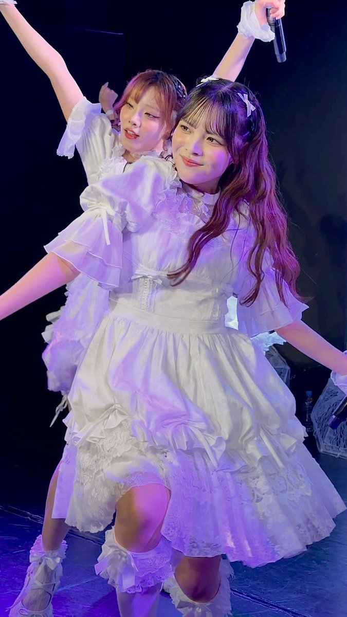 Two young women in frilly white dresses pose together on stage; one holds a microphone while raising her arm.