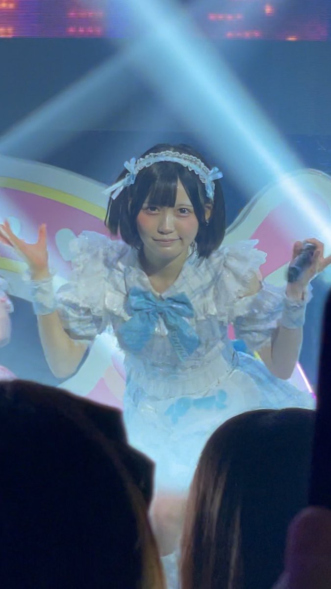 A young woman with short brown hair and a blue and white frilly maid-style outfit performs on a brightly lit stage, holding a microphone while making a playful gesture with her hands.